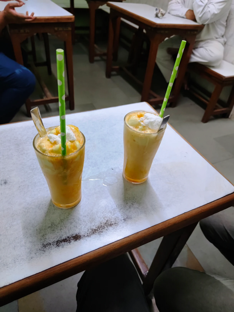 Tall glass of mango mastani topped with whipped cream and chopped nuts