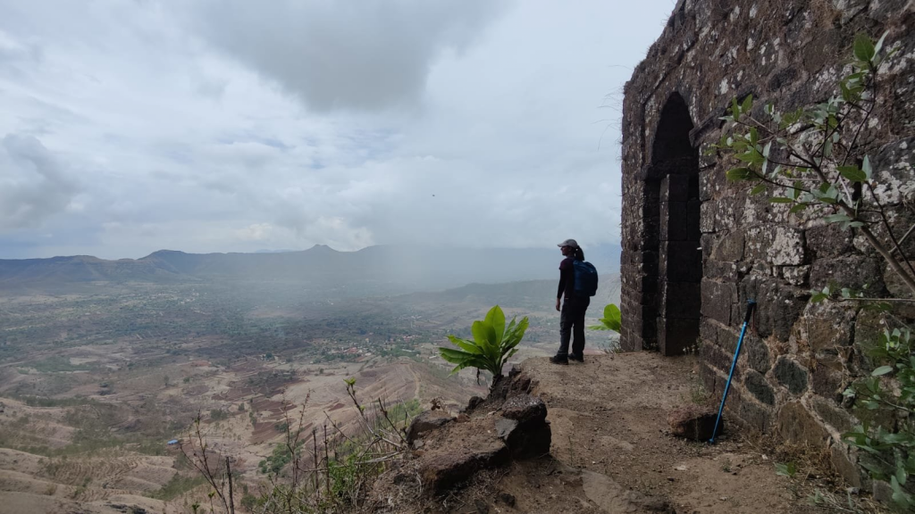 Weathered bastion walls and rock‑cut water cisterns clustered around an ancient temple, with seasonal waterfalls cascading along a grassy ridge