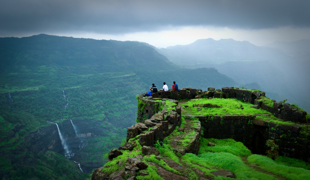 Ruins of Rajmachi Fort atop a forested hill, with trekkers gazing at the Sahyadri valley below.