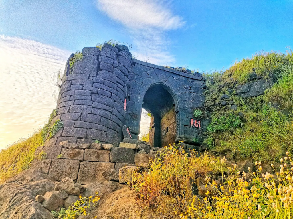 A massive stone gateway with Hanuman and Ganesh idols beside the “Eye of the Needle” rock aperture, set above lush valleys and the Amruteshwar Temple